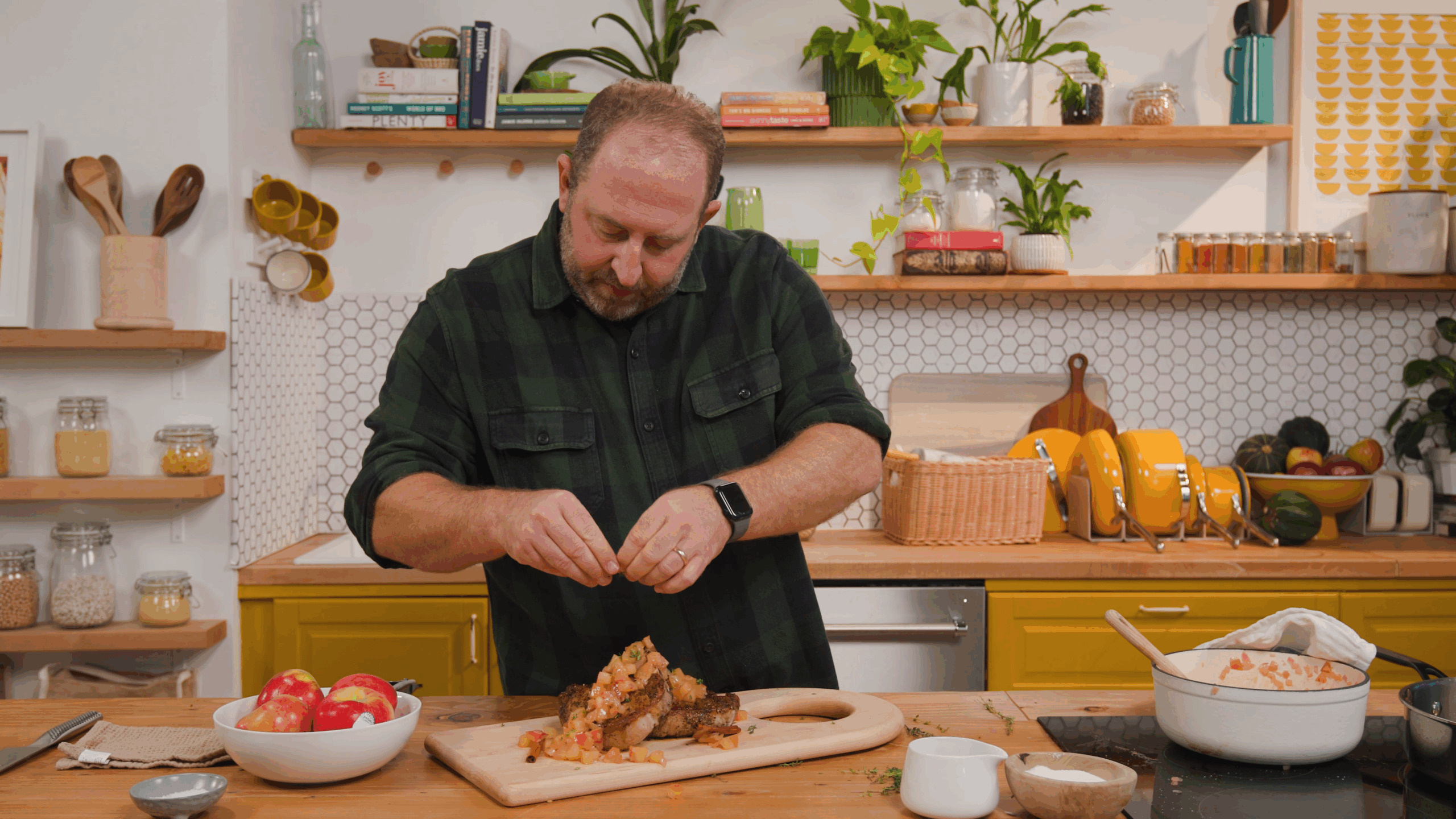 man sprinkling finishes on pork chops that have apple pieces on top in a bright kitchen with plants in the background