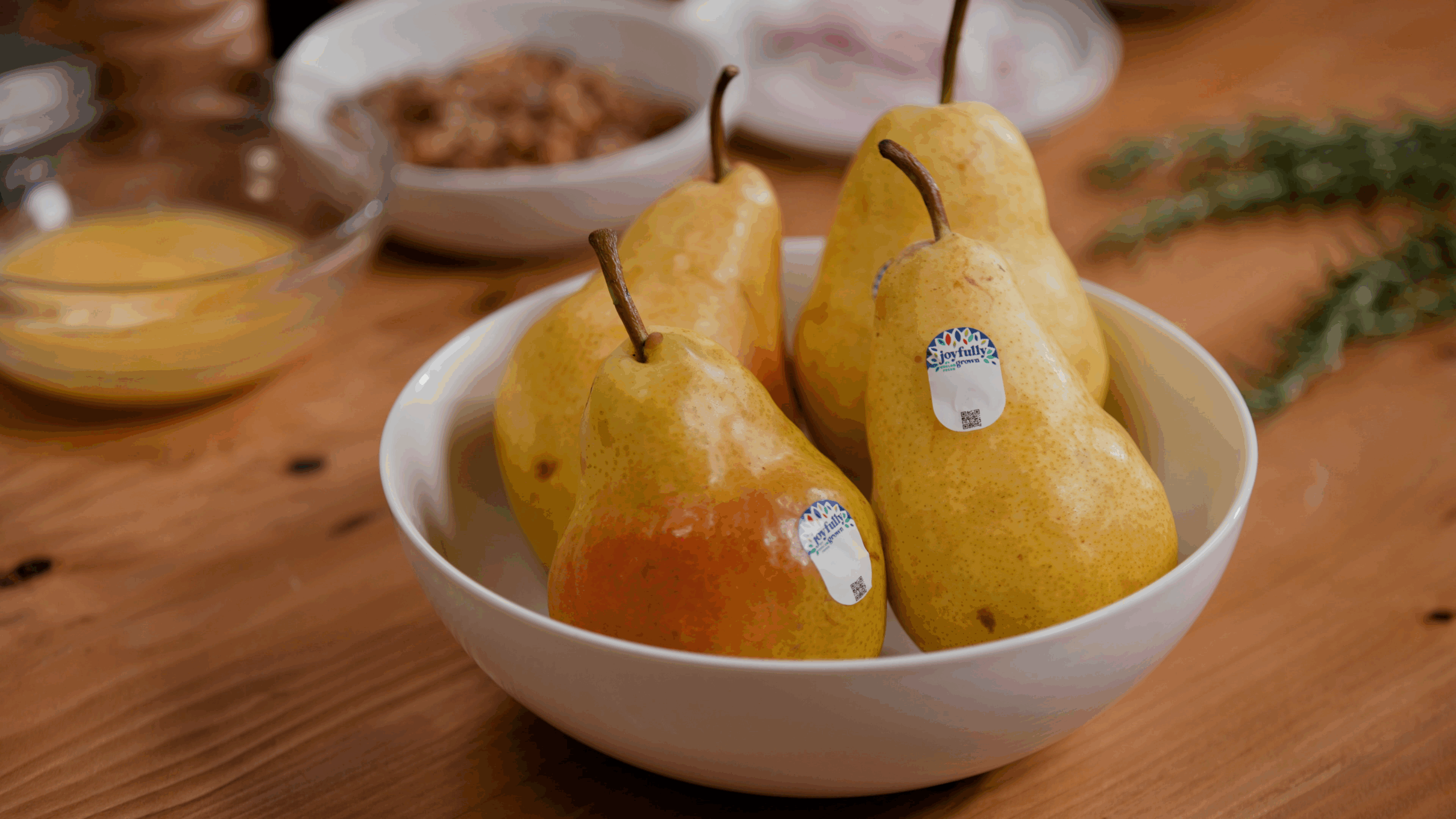 pears with Joyfully Grown stickers in a white bowl on a wood countertop with ingredients shown in the distant background